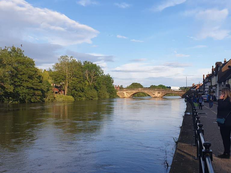 A shop filled street in Bewdley looking down the river Severn towards the tan stone, bridge which spans the river with 3 huge arches. The far side is tree covered with one red building in view.