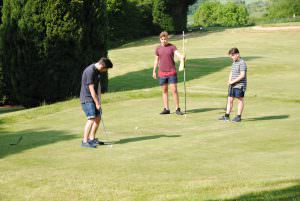 3 young teenagers playing pitch and put golf at Bank Farm. They are casually dressed in long shorts and tee shirts. One is about to put the ball on the green, the others stand and watch. The course is well mowed and is surrounded by tall green trees.