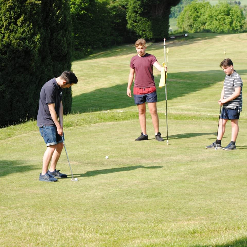 3 young teenagers playing pitch and put golf at Bank Farm. They are casually dressed in long shorts and tee shirts. One is about to put the ball on the green, the others stand and watch. The course is well mowed and is surrounded by tall green trees.