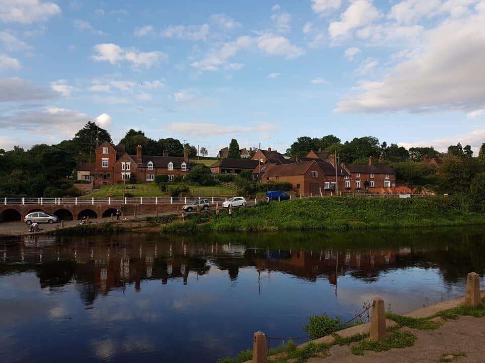 The view from Bank Farm of the small village of Arley is seen across the river Severn, the panorama is perfectly mirrored in the calm river’s waters. 4 cars are parked along the river’s edge and the few red brick houses with white windows gives a picture postcard impression.