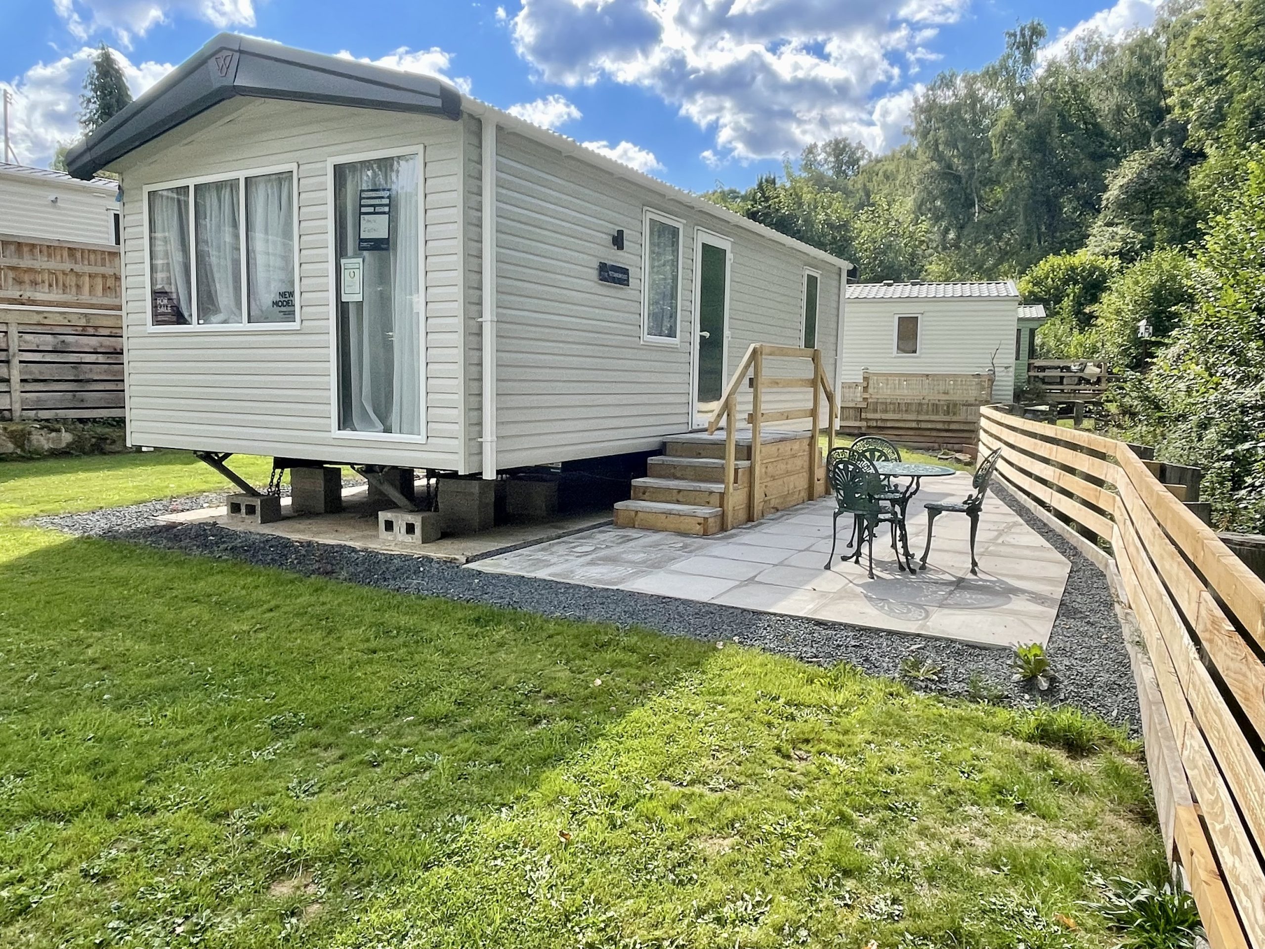 The side view of one of three Victory Stonewoods on site for sale. This static caravan has steps to the side entrance and a patio area with table and 4 chairs. its next to a small babbling brook.